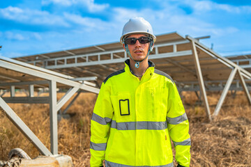 Smiling solar energy engineer in a reflective jacket and helmet stands confidently with arms crossed at a solar farm. Photovoltaic panels are visible in the background under a clear blue sky.