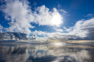 Obraz premium Serene coastal scene with a vibrant blue sky, fluffy clouds, sunlit reflections on wet sand, and calm ocean waves meeting a tranquil horizon.