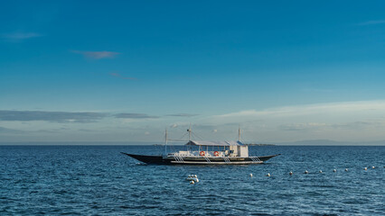 A beautiful traditional double-outrigger dugout bangka boat is anchored in the blue ocean. Life buoys on the railing. The awning over the deck. Ripples on the water. azure sky, clouds. Philippines.