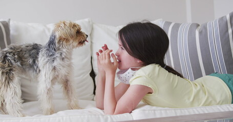 Caucasian girl spending time at home, lying on sofa with face mask, playing with pet dog, in slow mo