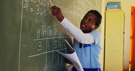 Side view close up of a young African schoolgirl standing at the front of the class reading from a b