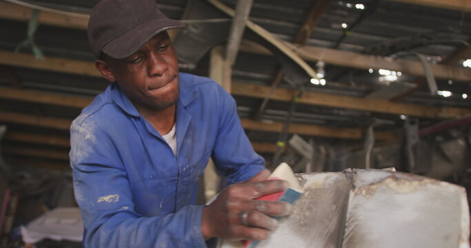 Front view of an African male panel beater in a township workshop, sanding and preparing surface of