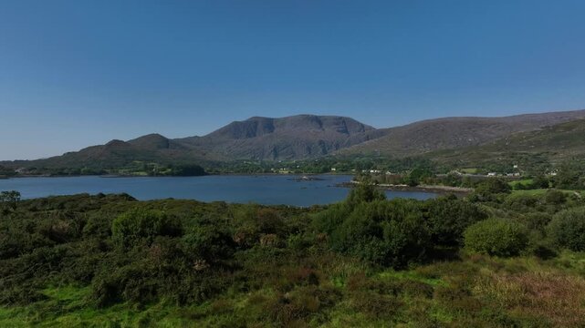 Adrigole Harbour, Beara Peninsula, County Cork, Ireland, September 2024. Drone pushes forwards over the wild rugged landscape towards the coast with the Caha Mountains in the distance under a blue sky