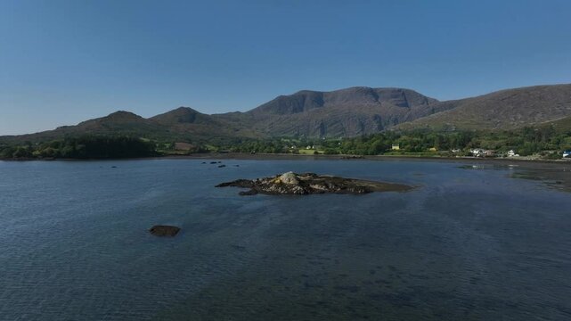 Adrigole Harbour, Beara Peninsula, County Cork, Ireland, September 2024. Drone pushes west low over the water with a stunning view of the Coomgira and Caha Mountains in the distance under a blue sky.