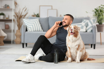 Sporty young man with Australian Shepherd dog talking by mobile phone on mat at home