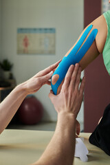 Close-up of therapist carefully applying blue sports tape on patient's arm during treatment session...
