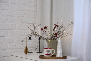 Hot cacao with marshmellows with Christmas dry tree branches with blurred lights and red berries, christmas tree on table in living room.