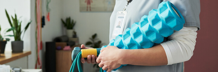 Close-up of healthcare professional holding rehabilitation equipment in clinic setting. Detail of hands and equipment, with plants and medical items in background
