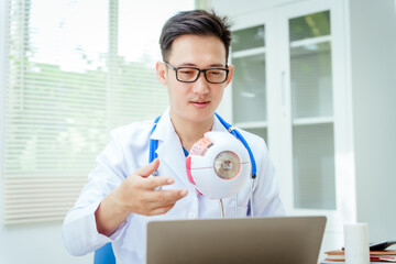 A male doctor wearing glasses sits at a desk in a hospital, explaining eye diseases like glaucoma,...