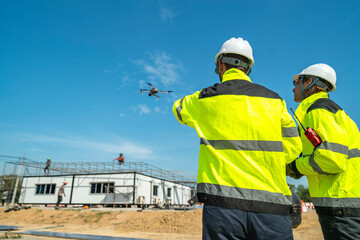Two engineers in high visibility jackets and helmets observing a drone flying over a construction...