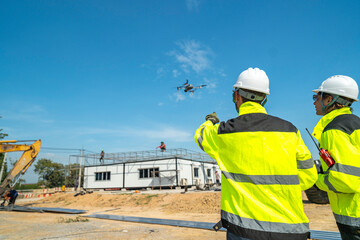 Two engineers in high visibility jackets and helmets observing a drone flying over a construction...