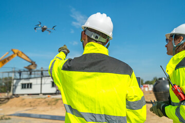 Fototapeta premium Two engineers in high visibility jackets and helmets observing a drone flying over a construction site, with machinery and modular buildings, highlighting technology and site monitoring.