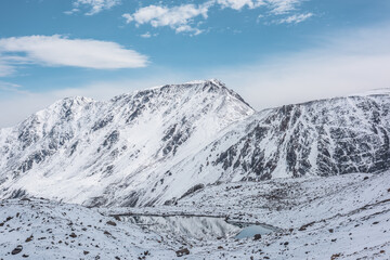 Cold snow-white landscape of snowy valley with alpine lake against snow-covered rocky mountain range under clouds in blue sky. Glacial lake, pure white snow and stones in high mountains in sunny day.