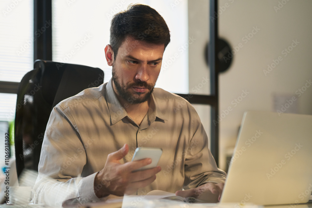 Poster serious male analyst looking at laptop screen and using mobile phone while watching online video or 