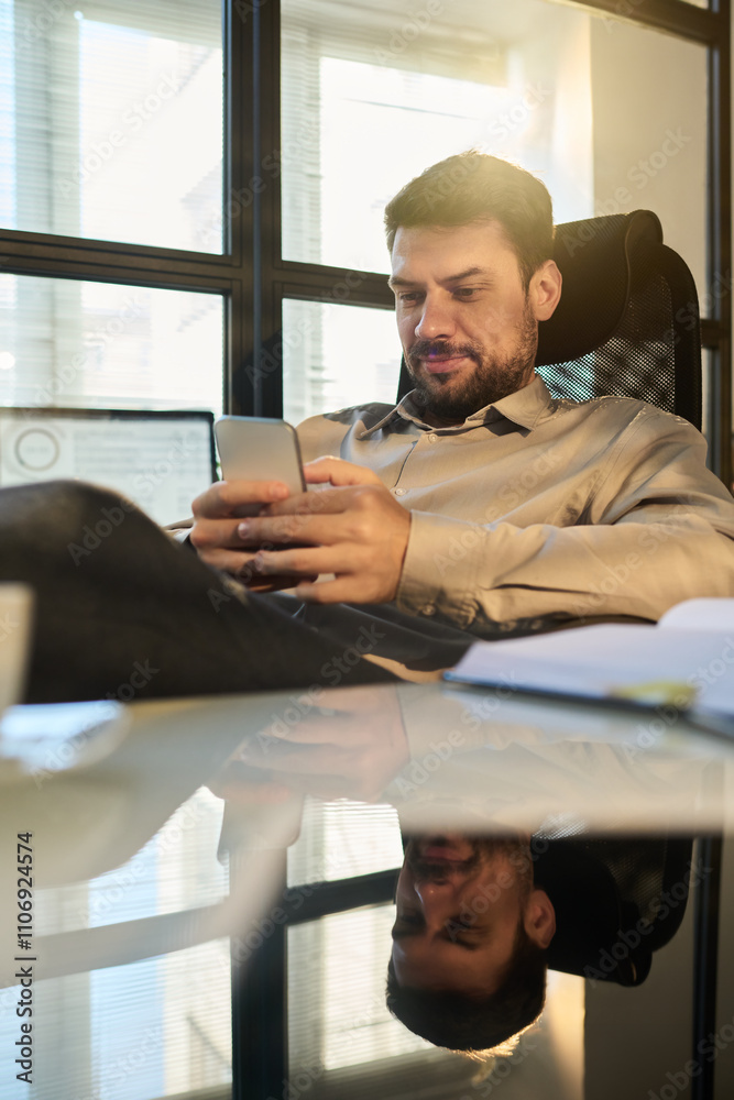 Poster Young restful office manager in smart casualwear texting in smartphone while enjoying break by workplace with notepad