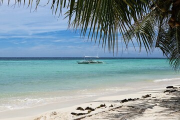 A serene beach scene features a bangka boat floating on clear turquoise waters, framed by palm leaves. The sandy shore is calm, with gentle waves lapping at the beach.