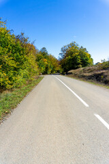 An asphalt road with a white dividing line passes through trees and bushes with autumn leaves. Hills and clouds are in the background