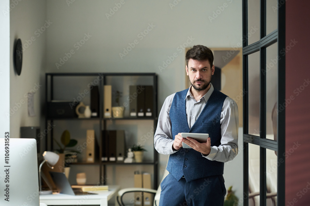 Poster Young serious businessman using tablet and looking at camera while standing in coworking space with desks and shelves