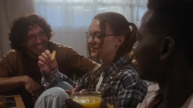 Group of young African American and Caucasian male and female friends sitting at coffee table in living room, talking, eating snacks and drinking juice