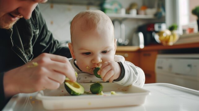 Man is feeding a baby avocado slices. The baby is eating the avocado slices and the man is helping him