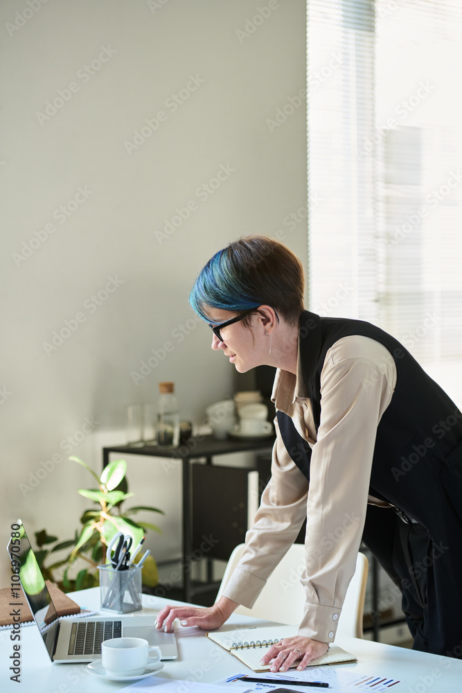 Poster side view of young female ceo bending over workplace with laptop and looking at device screen while 