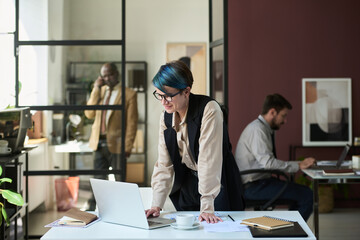 Young smiling female broker or analyst standing by workplace in coworking space and looking at...