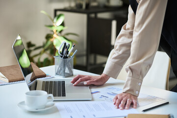 Hands of young businesswoman in smart casualwear bending over desk and using laptop while analyzing...