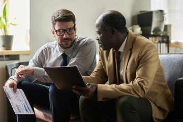 Two intercultural male analysts or economists sitting on sofa in office and discussing financial data in document at meeting
