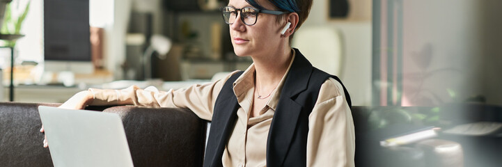 Fototapeta premium Header of young serious female analyst with laptop on her knees sitting on leather couch in office and working with online data