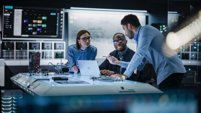 Diverse Team of Engineers Collaborating on Manufacturing Project in a Research Facility. Colleagues Analysing Data Using Laptop Computer and Brainstorming for a Technologically Advanced Invention