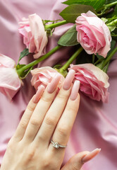 Woman hand with pink long nails is showing engagement ring with pink roses on silk background