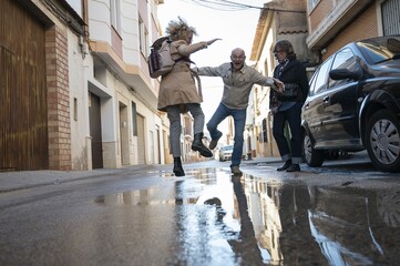 playful middle-aged adult friends jumping in a puddle on a narrow street in a small town 
