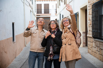 Cheerful senior friends standing on a residential street, looking up and waving excitedly, radiating happiness and camaraderie during a pleasant outing