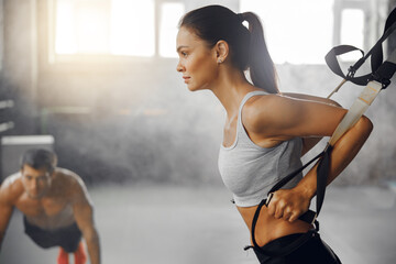 A woman performs suspension training while a man works out in the background, showcasing gym energy.