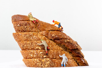 Miniature figurines climbing a stack of multigrain bread slices, resembling a rugged mountain. A whimsical and playful concept for food and adventure themes.