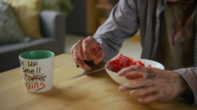 Medium closeup of hungry bloodied undead guy with decomposing flesh eating lunch at home, spicing bowl of raw meat or brains with pepper grinder, dropping spoon on table before putting food in mouth