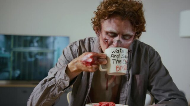 Medium closeup of young male living corpse with white eyes sitting at breakfast table with bowl of raw brains, lifting mug with dark humor print with shaky hand and drinking coffee. Zombie movie on TV
