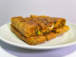 A platter of golden-brown martabak telur, a traditional Indonesian street food, is presented on a white plate. Each slice reveals a savory filling of egg, meat, chives, and other ingredients