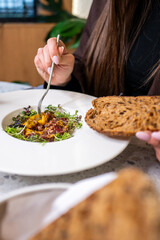 A close-up of a hand holding a fork, poised to eat salad. A plate of fresh greens garnished with colorful ingredients is centered, with a slice of whole grain bread visible in the foreground.