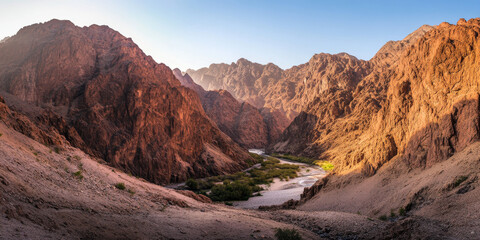 A dramatic desert canyon with towering red rock formations and a winding river at the bottom. The warm sunlight highlights the rugged beauty of the landscape.