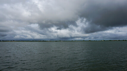 A vast water body under a dramatic cloudy sky, with reflections and a distant green shoreline. The...