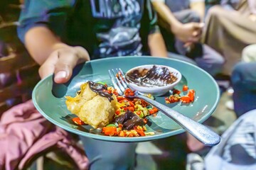 A person's hand holding a green leaf cup filled with a chocolate sauce, red chili peppers, crispy fried tofu, and a fork.