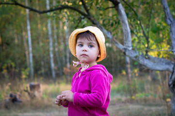 Little girl or child with sad expression wearing panama or headdress and pink sweater on walk in park or village. Blurred background with trees or forest. Portrait of child