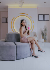 Obraz premium Portrait of smiling asian woman looking at camera holding skincare cream in both hands sitting on the couch at home. Beauty concept, product, reviews and advertisement.