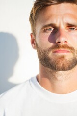 Fototapeta premium A rugged young man with a well-groomed beard and short hair, wearing a white shirt, partially lit by bright sunlight, creating a dramatic shadow effect on a minimalist white background