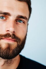 Obraz premium A close-up of a young man with piercing blue eyes and a neatly groomed beard, wearing a dark shirt, captured against a soft blue flat background