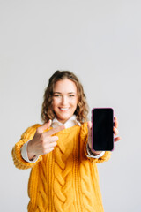 Naklejka premium Smiling young woman pointing at mobile phone screen, presenting a new app. Holding a smartphone with a blank display, isolated on white background. Ideal for app promotion and tech ads.