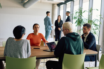 A diverse team of business professionals engaged in a discussion around a conference table in an office, while their colleagues collaborate in the background