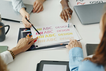 Group of diverse colleagues brainstorming creative design concepts in professional setting Working hands pointing at colorful print samples on worktable