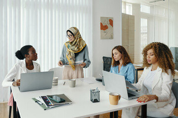 Group of diverse women engaged in business discussion around meeting table in modern office. Each...